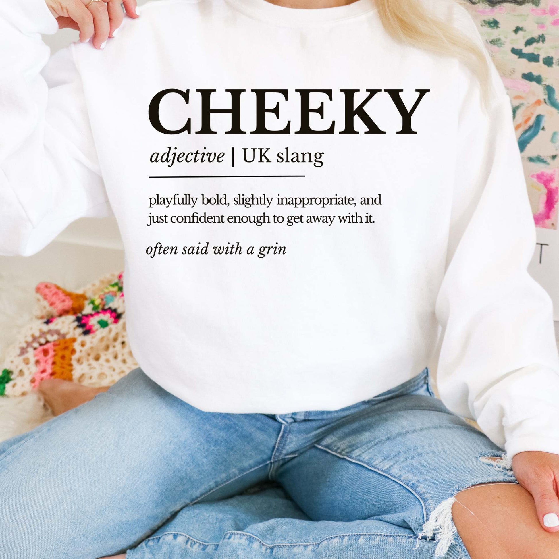 Person wearing a white sweatshirt with 'CHEEKY' definition text, sitting on a colorful couch.