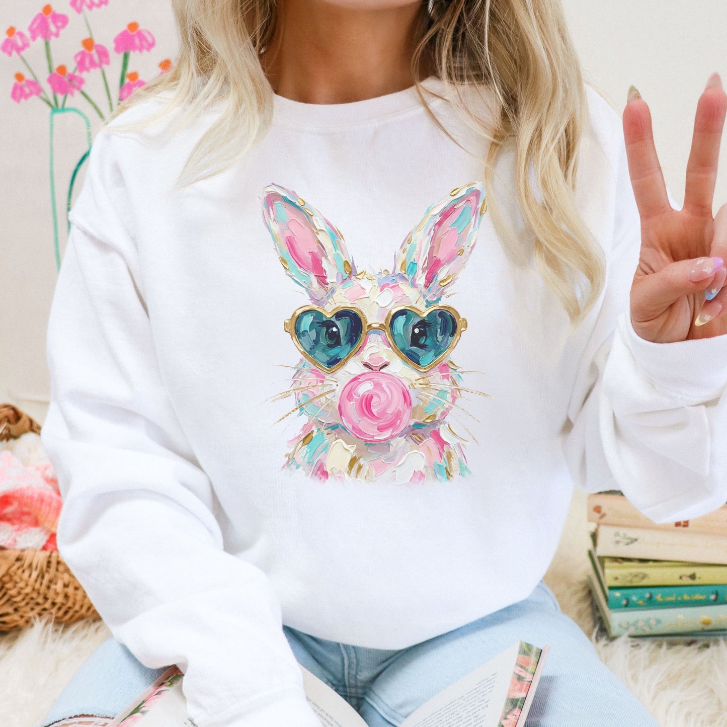 Person wearing a white sweatshirt with a colorful bunny design, sitting on a couch with books and flowers in the background.
