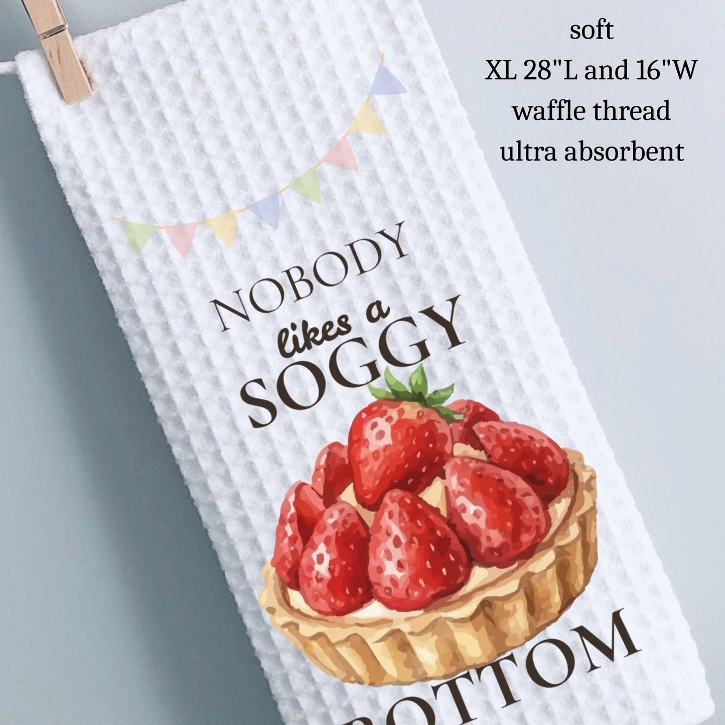 Waffle-textured towel with strawberry design and text, hanging on a clothespin against a light gray background.