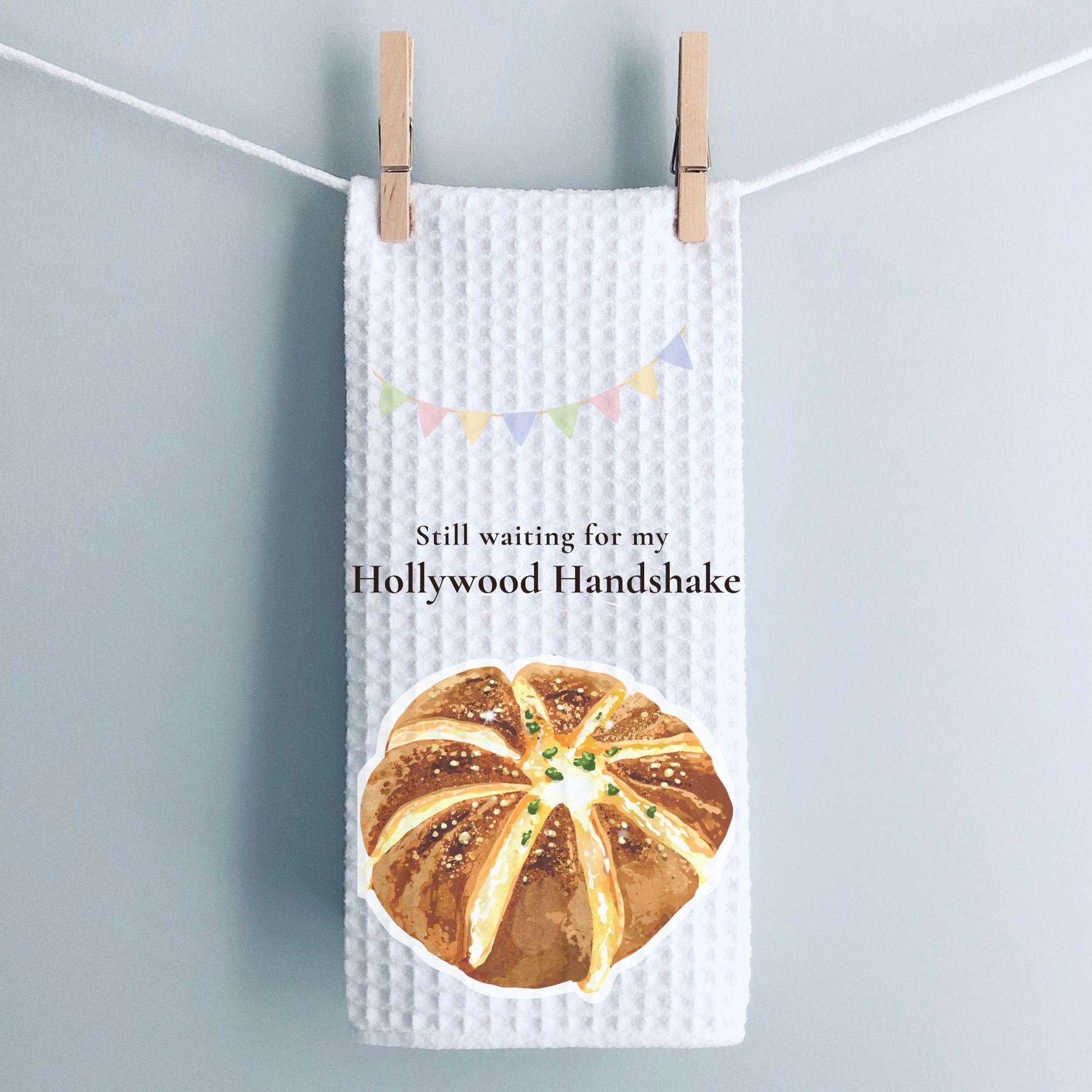 Tea towel with bread design and text 'Still waiting for my Hollywood Handshake' hanging on a line.