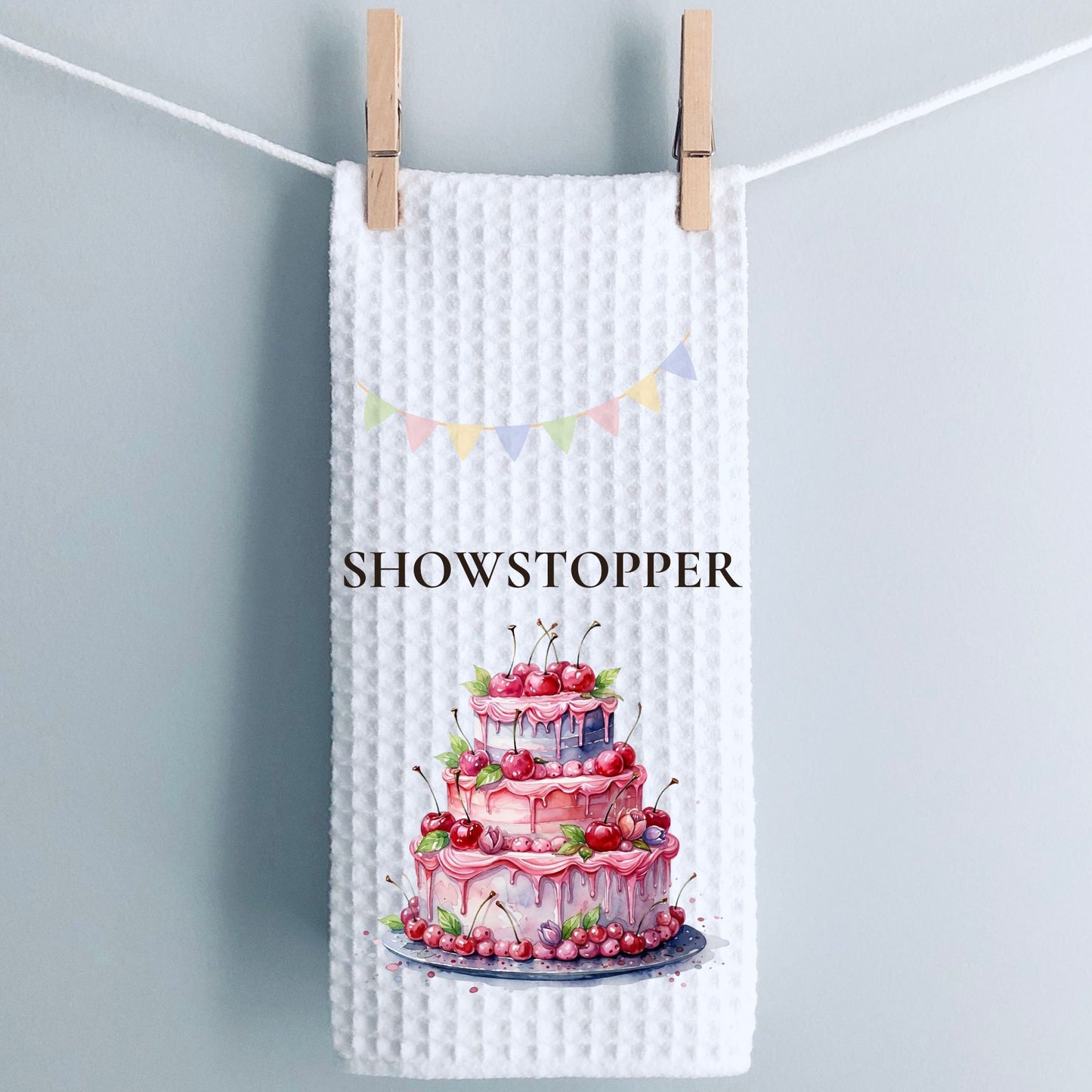 White textured towel with a cake illustration and 'SHOWSTOPPER' text, hanging on a line.
