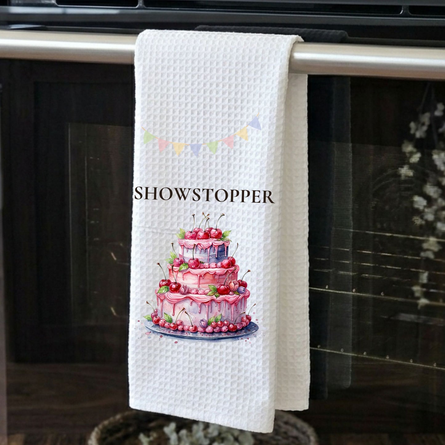 White towel with a cake design and 'SHOWSTOPPER' text hanging on a kitchen towel rack.