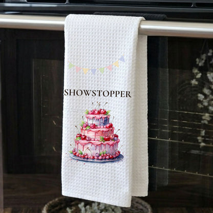 White towel with a cake design and 'SHOWSTOPPER' text hanging on a kitchen towel rack.