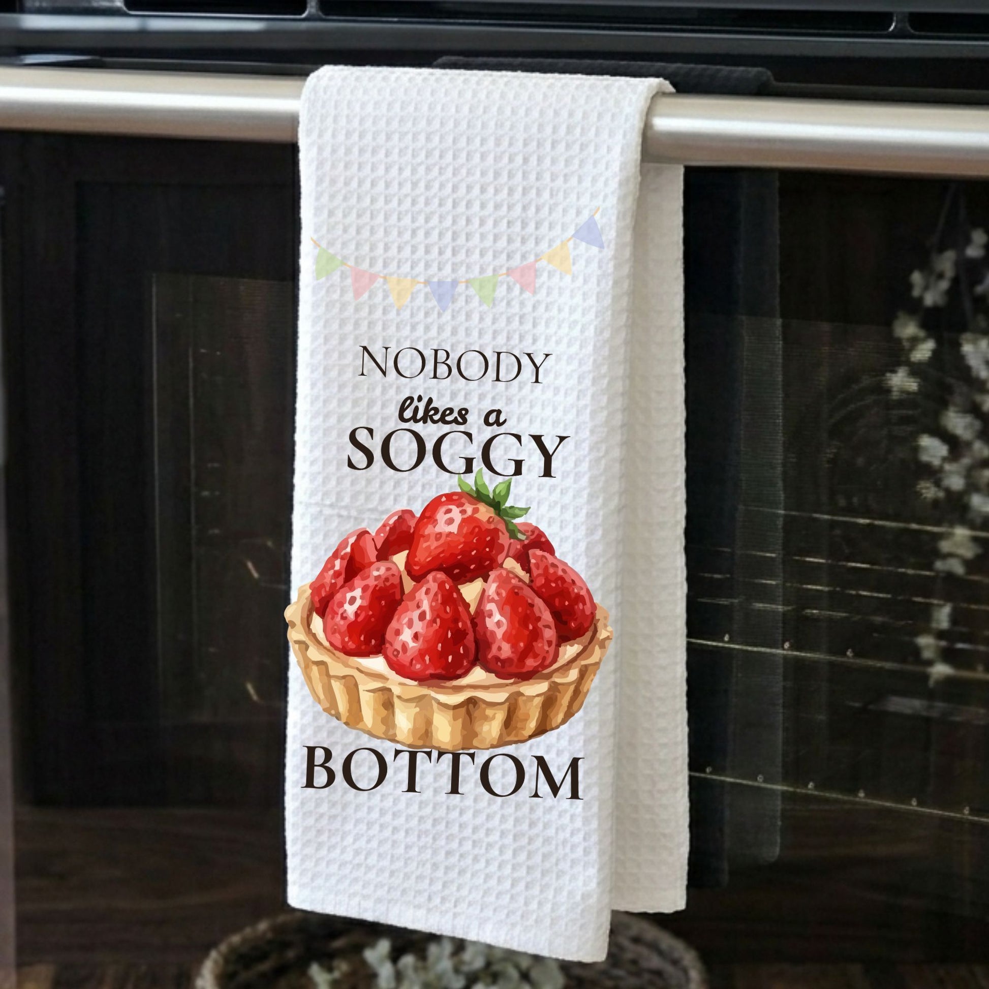 Kitchen towel with strawberry tart design and humorous text hanging on a stove handle.