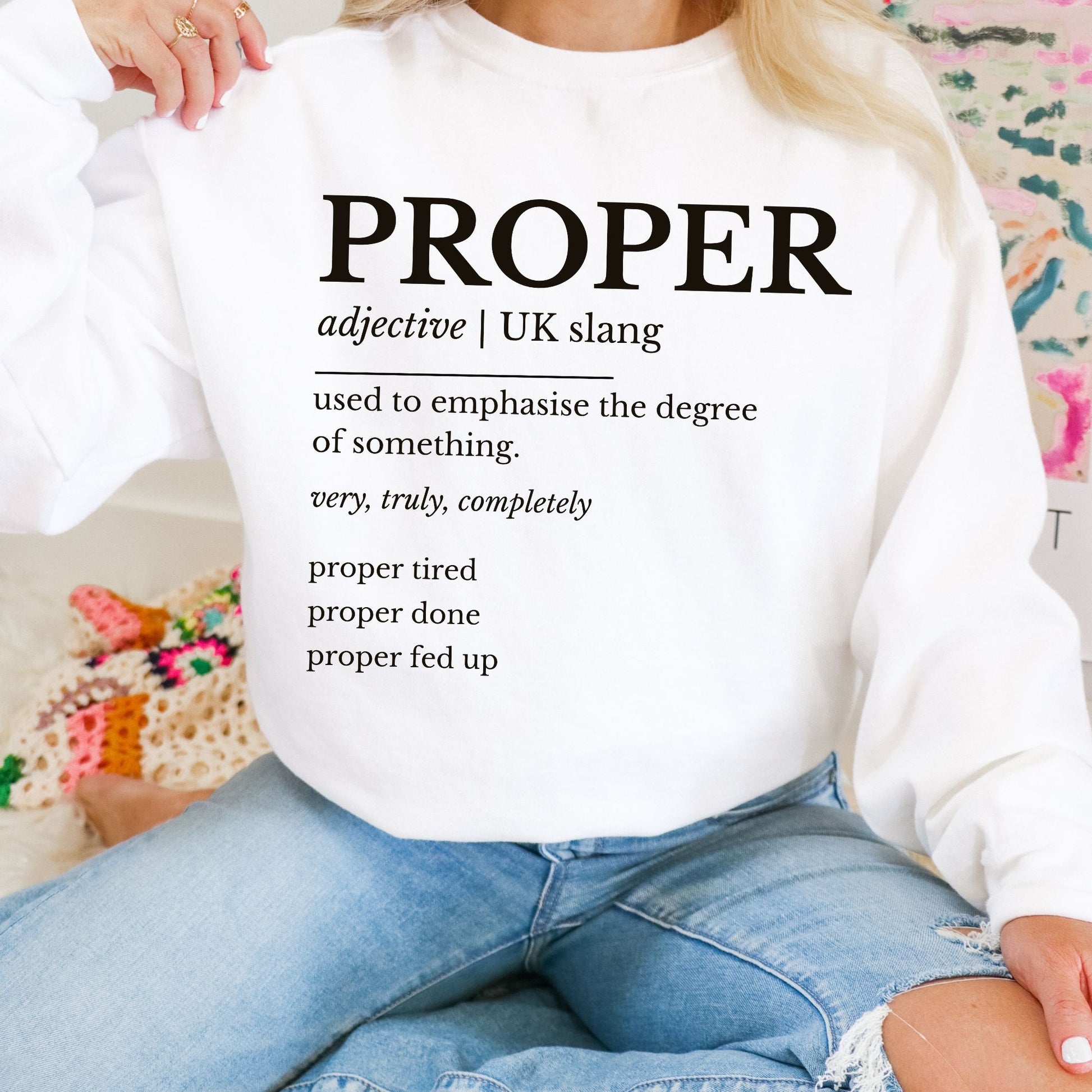 Person wearing a white sweatshirt with 'PROPER' definition text, sitting on a colorful couch.