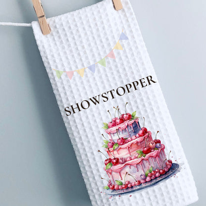 White towel with a cake illustration and 'SHOWSTOPPER' text, hanging on a line.